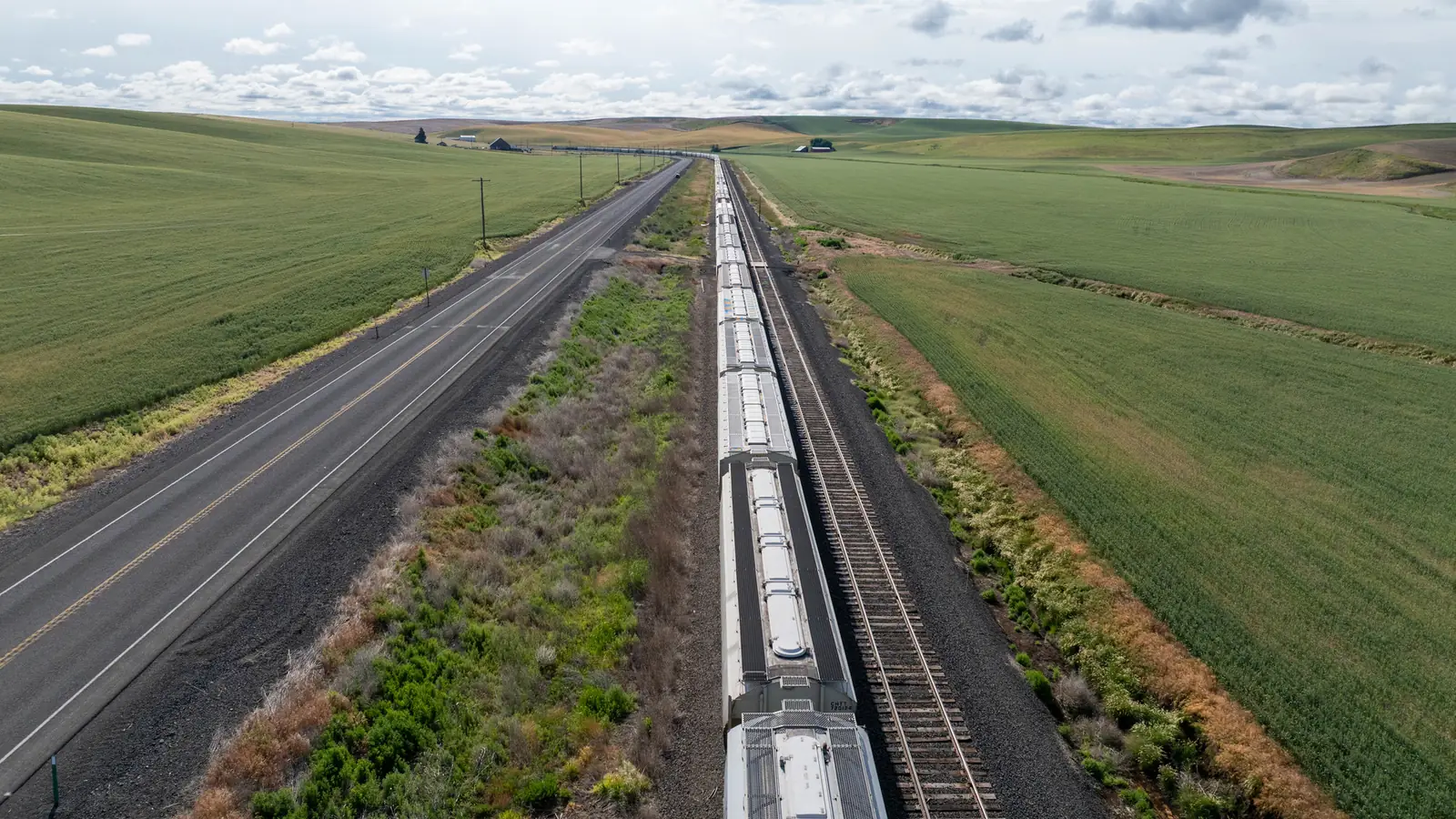 A freight train curves through farmland beside a highway, seen from a drone.